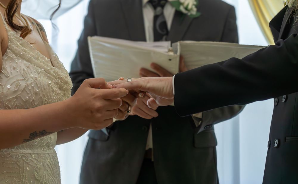 Bride and groom exchanging rings during wedding ceremony with officiant holding book.