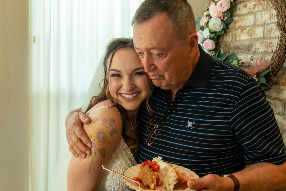 Smiling bride with floral tattoo shares a warm hug with older man holding wedding cake plate.