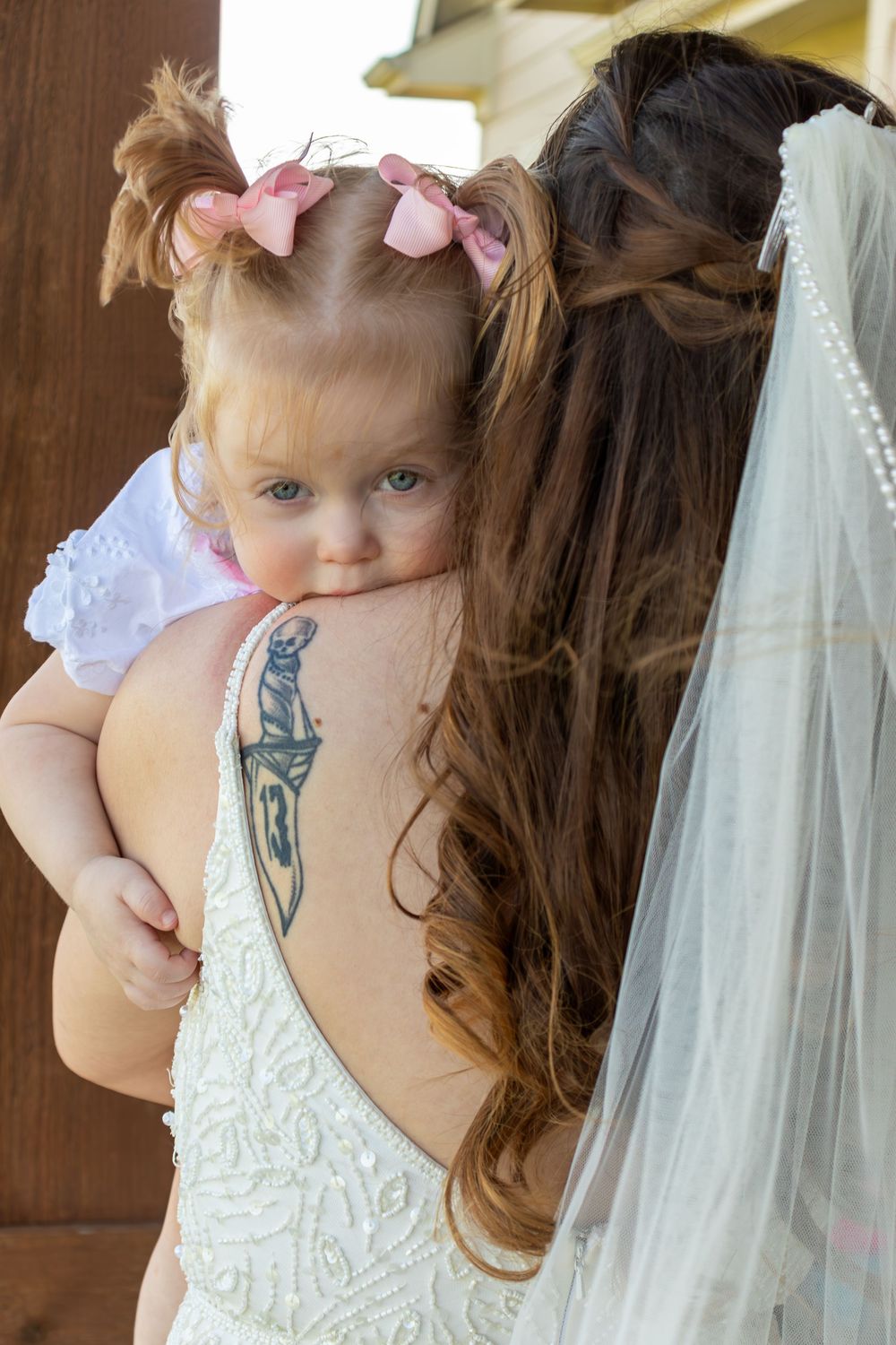 Bride in backless beaded wedding dress with tattoo holding young toddler girl with pink bows.