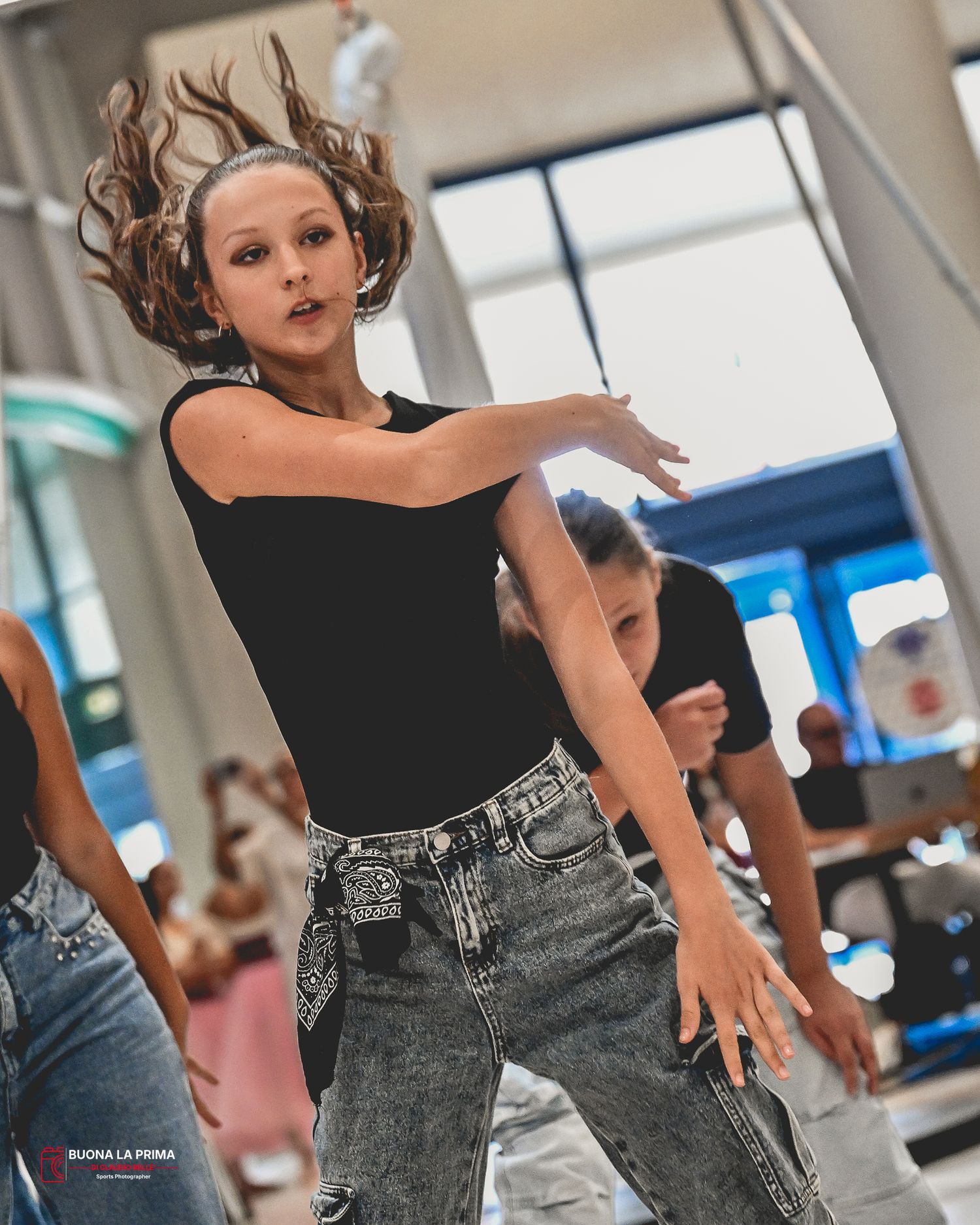 Young dancer in black top and jeans mid-performance, hair flying, captured in dynamic motion at a dance studio.