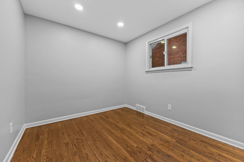 Empty room with warm hardwood floors, recessed lighting, and a small window revealing brick exterior.