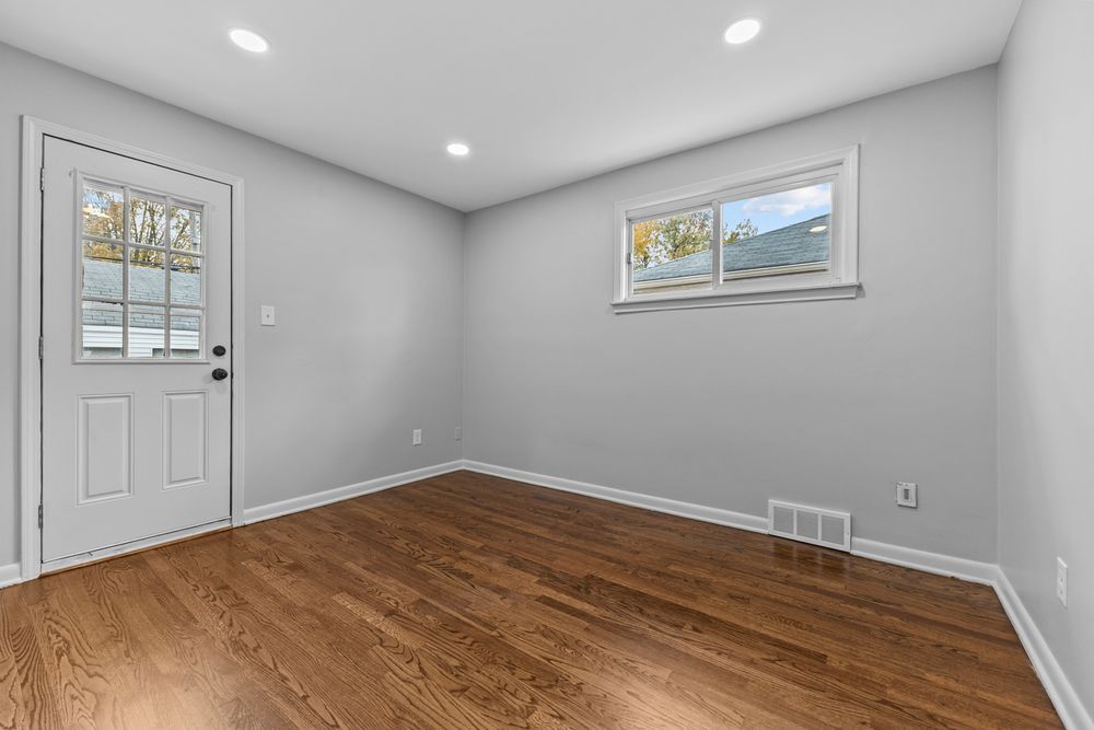Bright empty room with hardwood floors, recessed lighting, two windows, and a white exterior door.