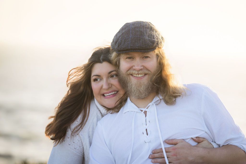 A smiling couple poses together on a beach at golden hour, the man wearing a flat cap and beard, the woman with long dark hair.