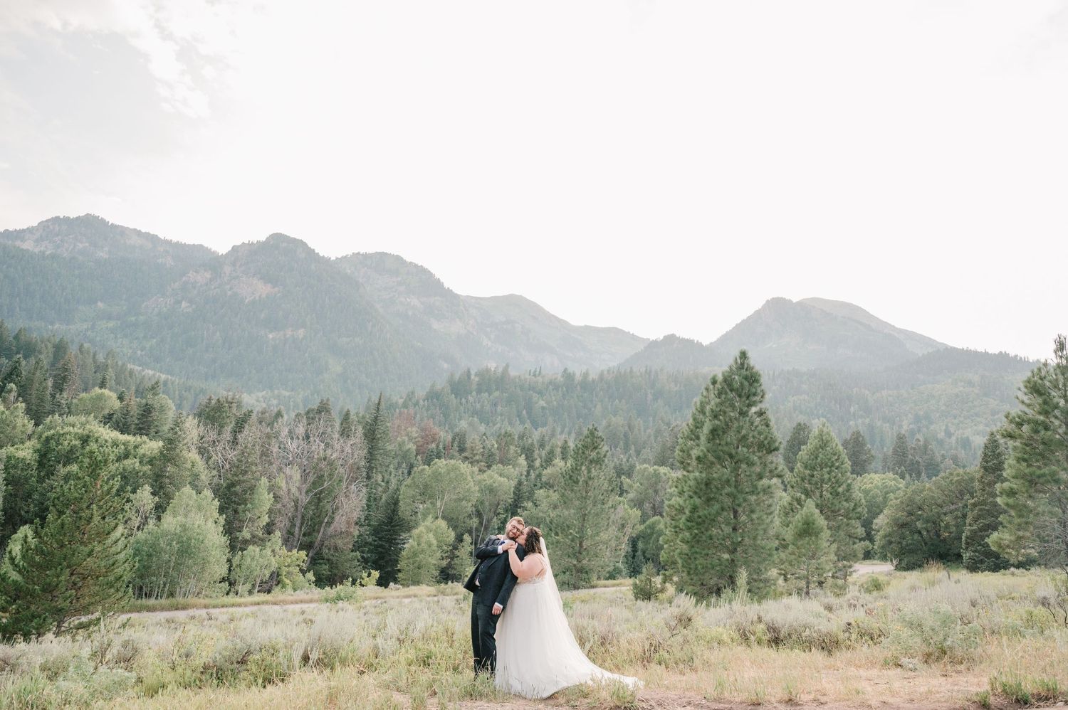 A couple embraces in their wedding attire against a scenic mountain landscape with evergreen trees in the background.