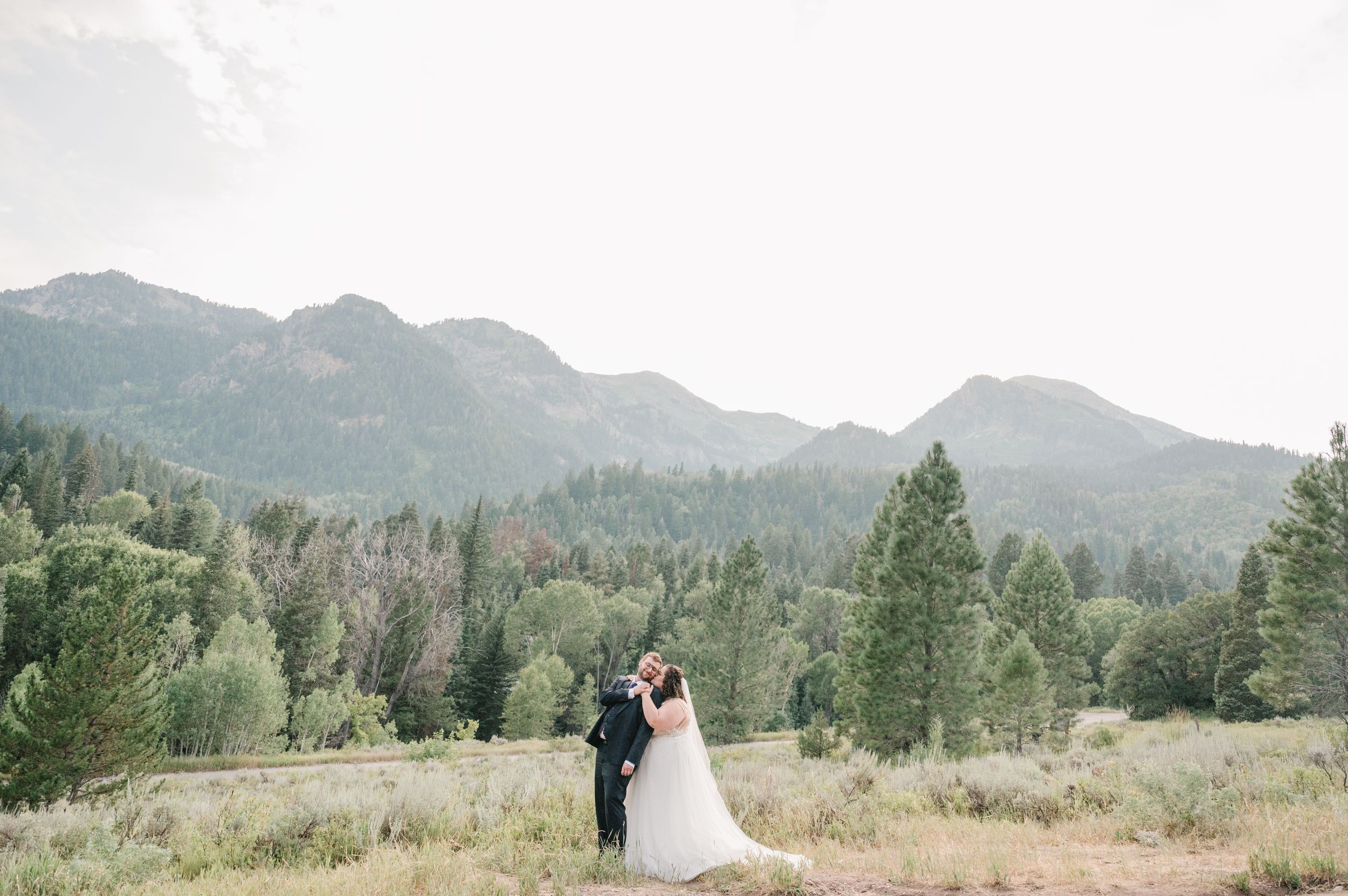 A couple embraces in their wedding attire against a scenic mountain landscape with evergreen trees in the background.
