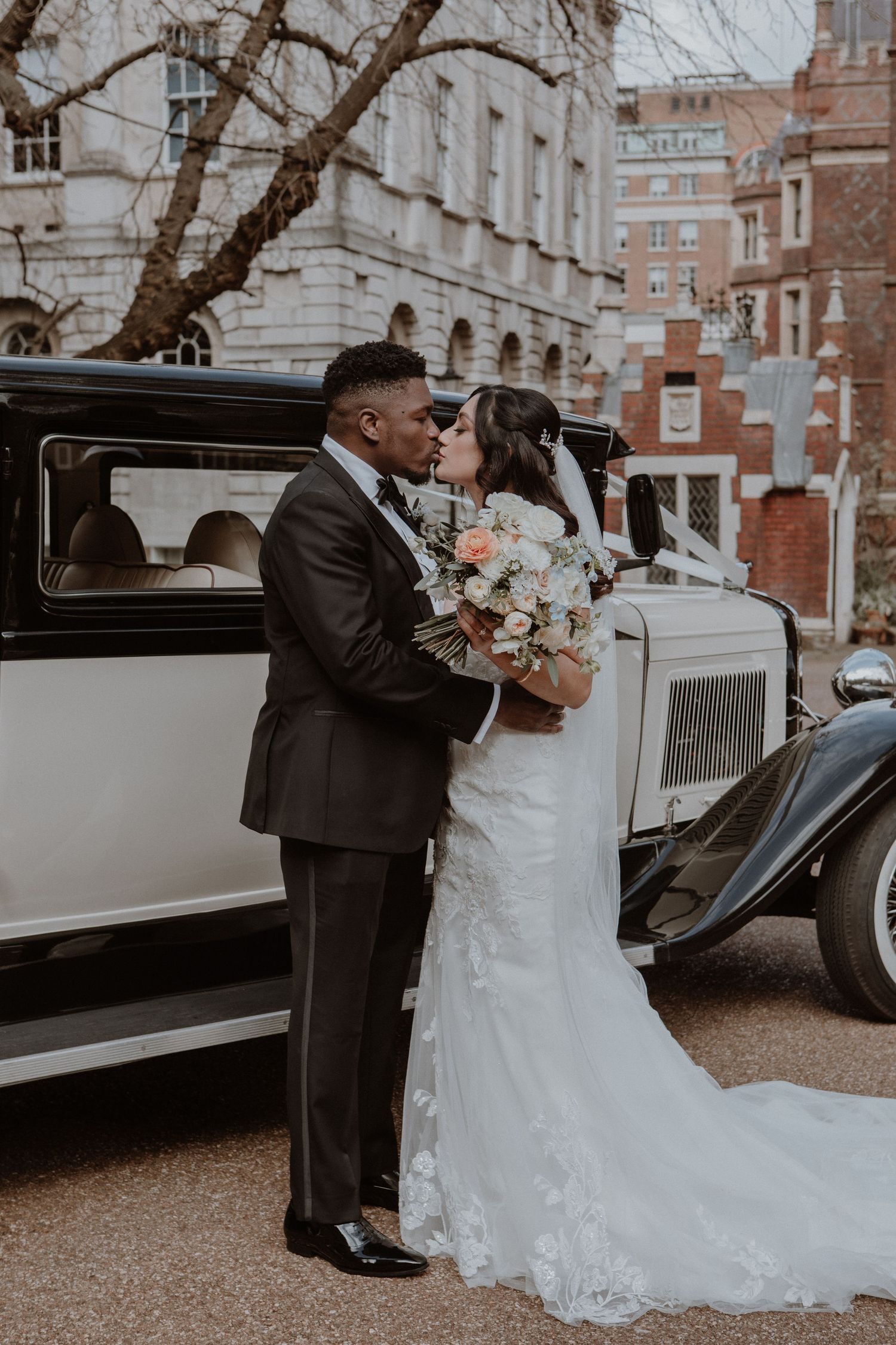 A newlywed couple embracing by a classic vintage car on their wedding day.
