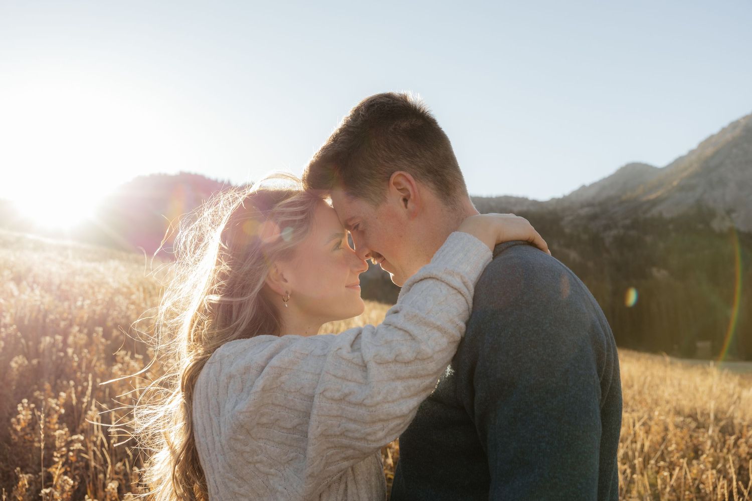 Romantic couple touching foreheads in golden sunlit field with mountains behind.