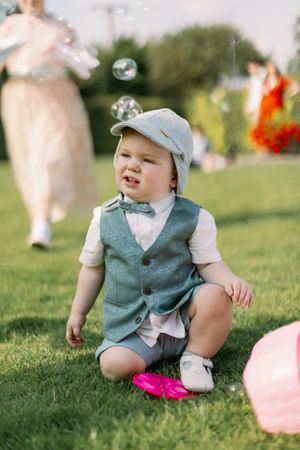 A toddler in a vest and cap sits on grass with bubbles floating overhead.