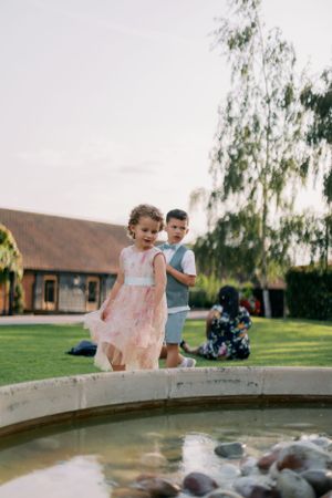 Children in dress clothes explore a stone fountain in a garden setting.