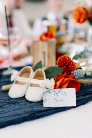 Wedding detail shot of white baby shoes and orange florals on navy blue fabric with a place card.
