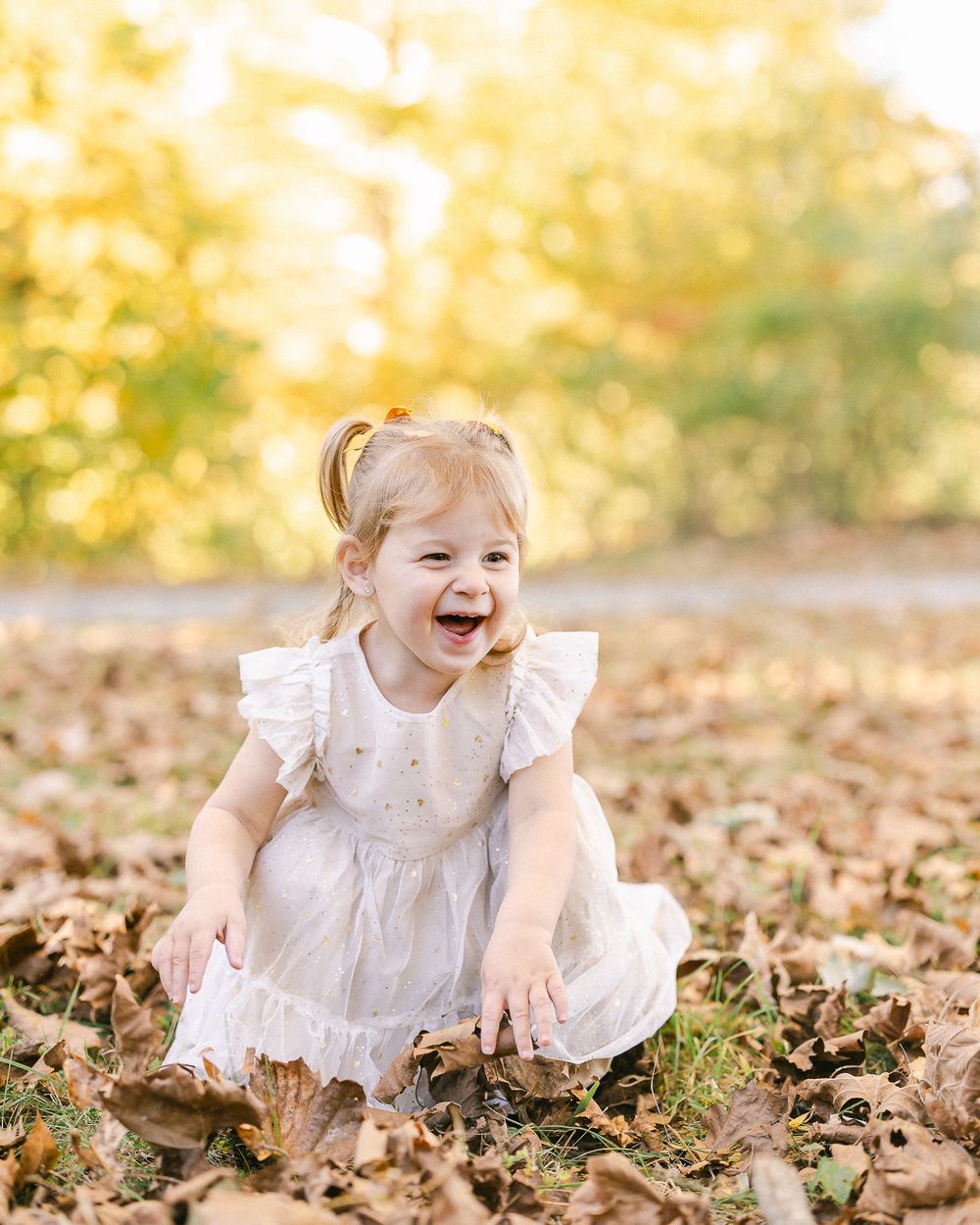 A child in a white dress laughs while playing in autumn leaves.