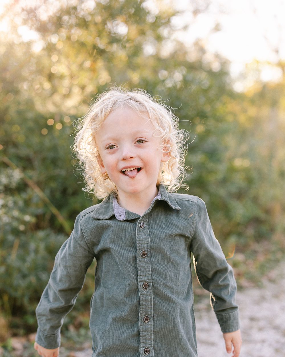 A child with curly blonde hair smiles brightly while wearing a button-down shirt outdoors.