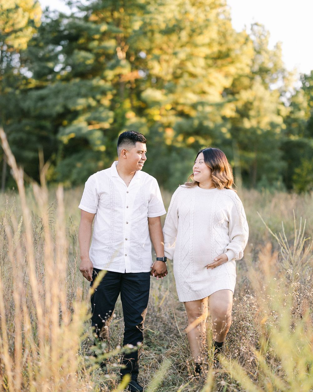 Expecting couple walking hand in hand through tall grass during golden hour.