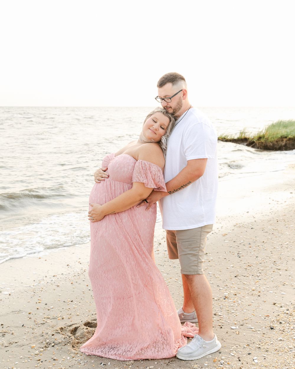 A loving couple embraces on the beach at sunset with the woman wearing a flowing pink dress.
