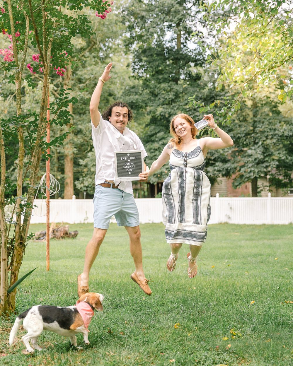 A couple jumps joyfully in their backyard with their dog while holding a sign.