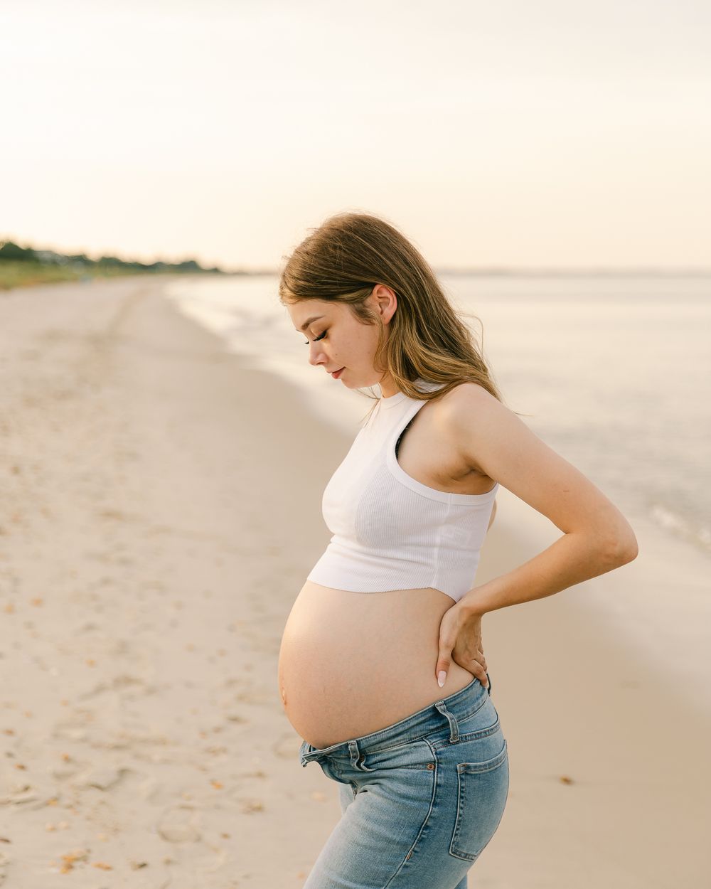 Pregnant woman in white tank top and jeans poses on beach at sunset.