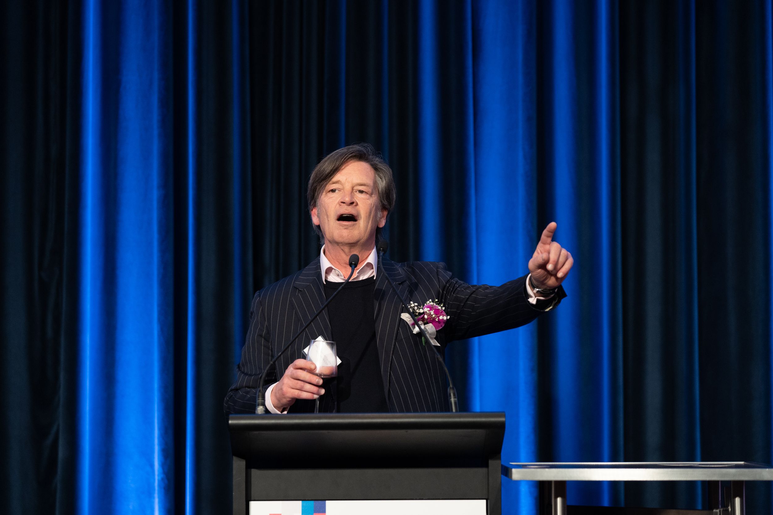 Speaker gestures enthusiastically while presenting at podium with blue curtain background.