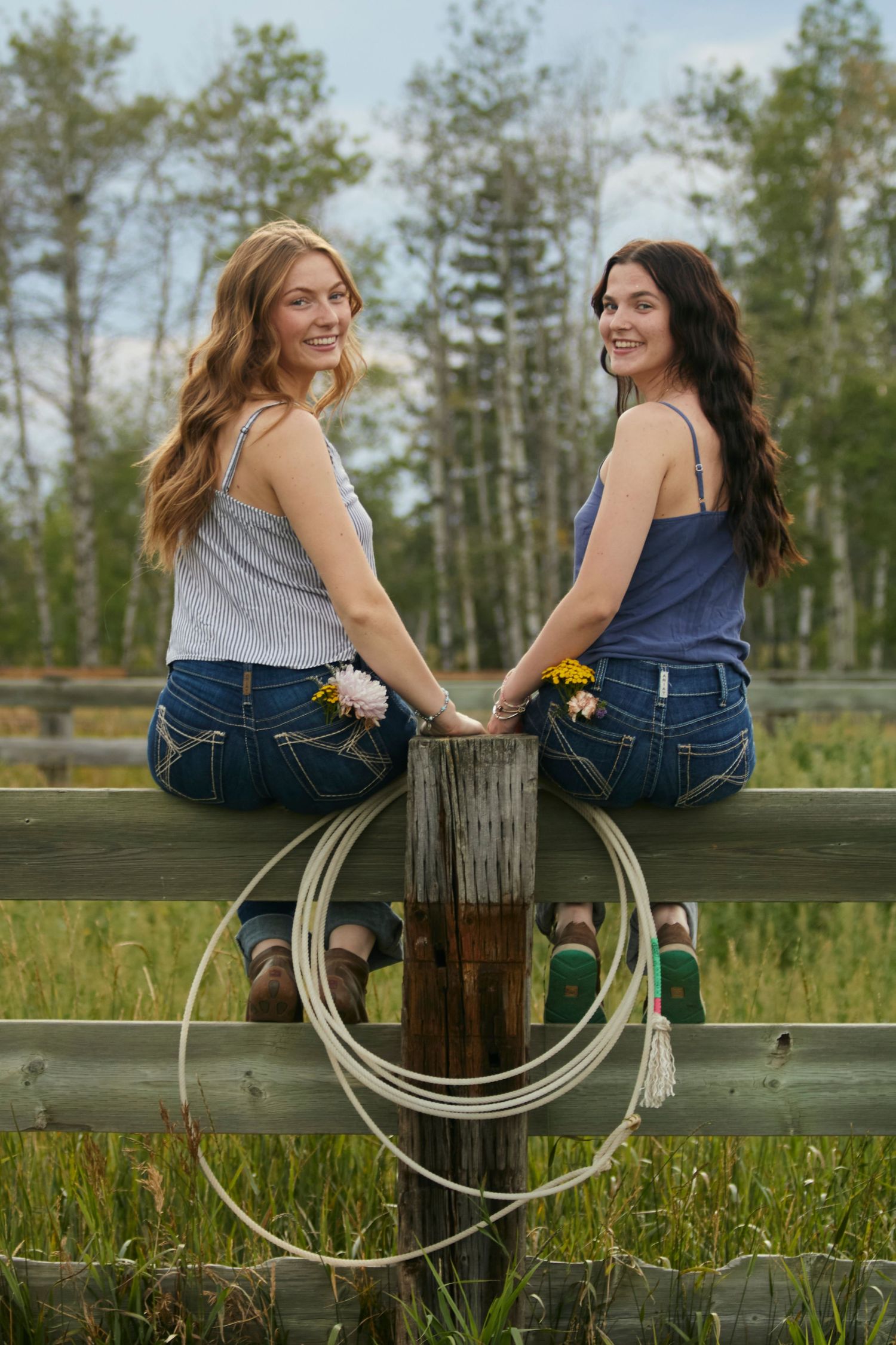 Two young women sitting on a wooden fence in jeans and tank tops, smiling in a rural field with flowers and a rope, summer countryside vibe.