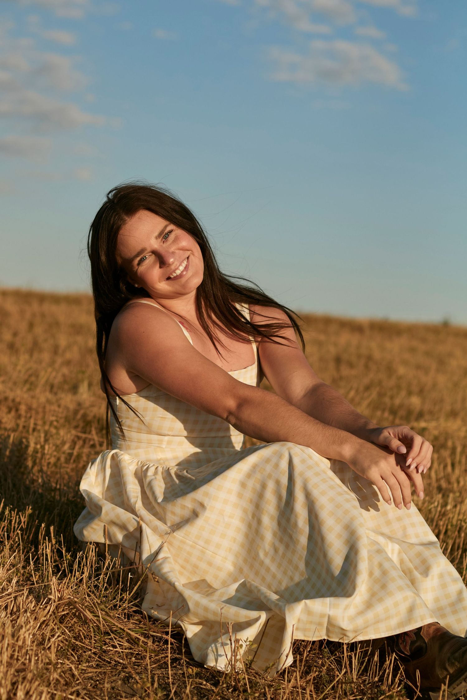 Woman sitting in a golden field at sunset, smiling in a light summer dress, capturing warm rustic and carefree countryside vibes.