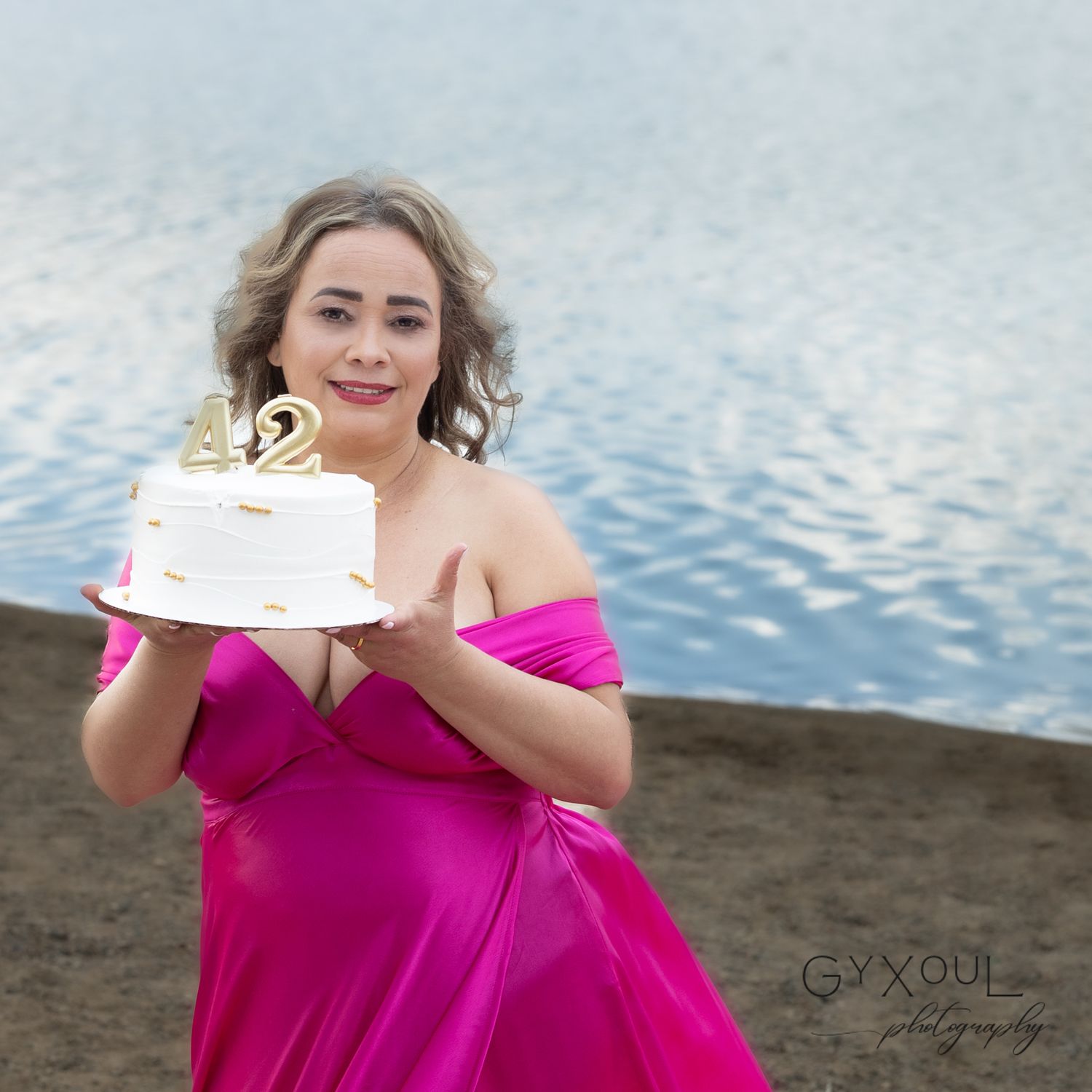 Person in pink formal dress holding small celebration cake by waterfront.