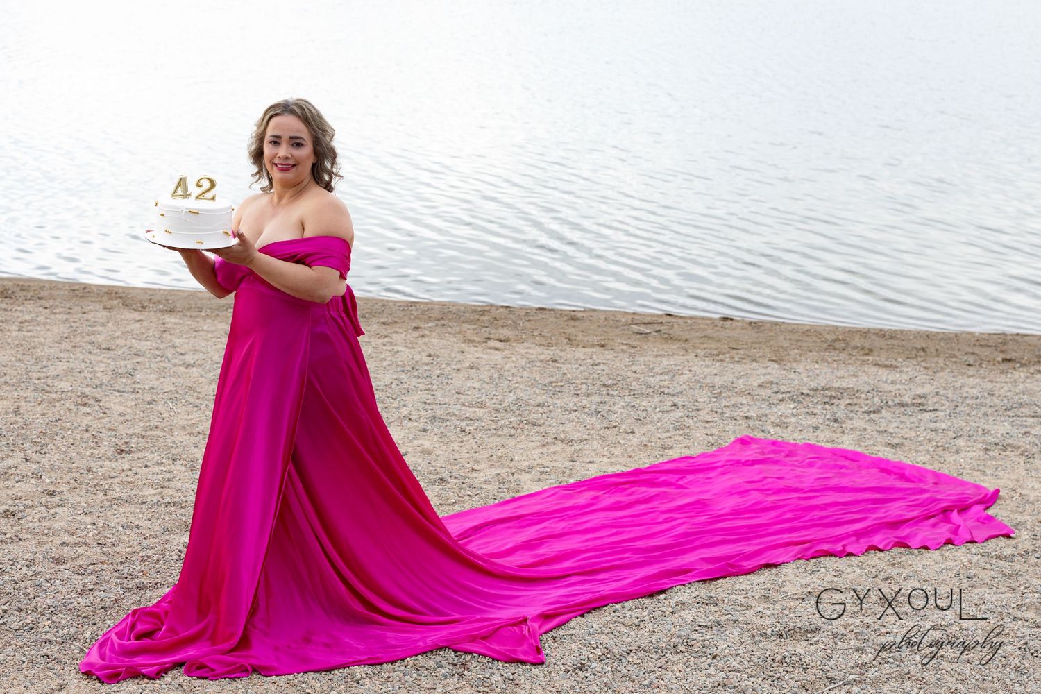 Person posing with flowing pink dress on beach holding celebration cake.