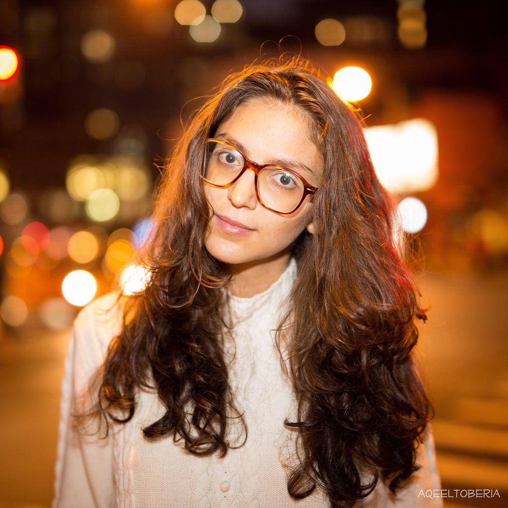 Portrait of someone with wavy dark hair and glasses in warm evening lighting with bokeh effects in the background.