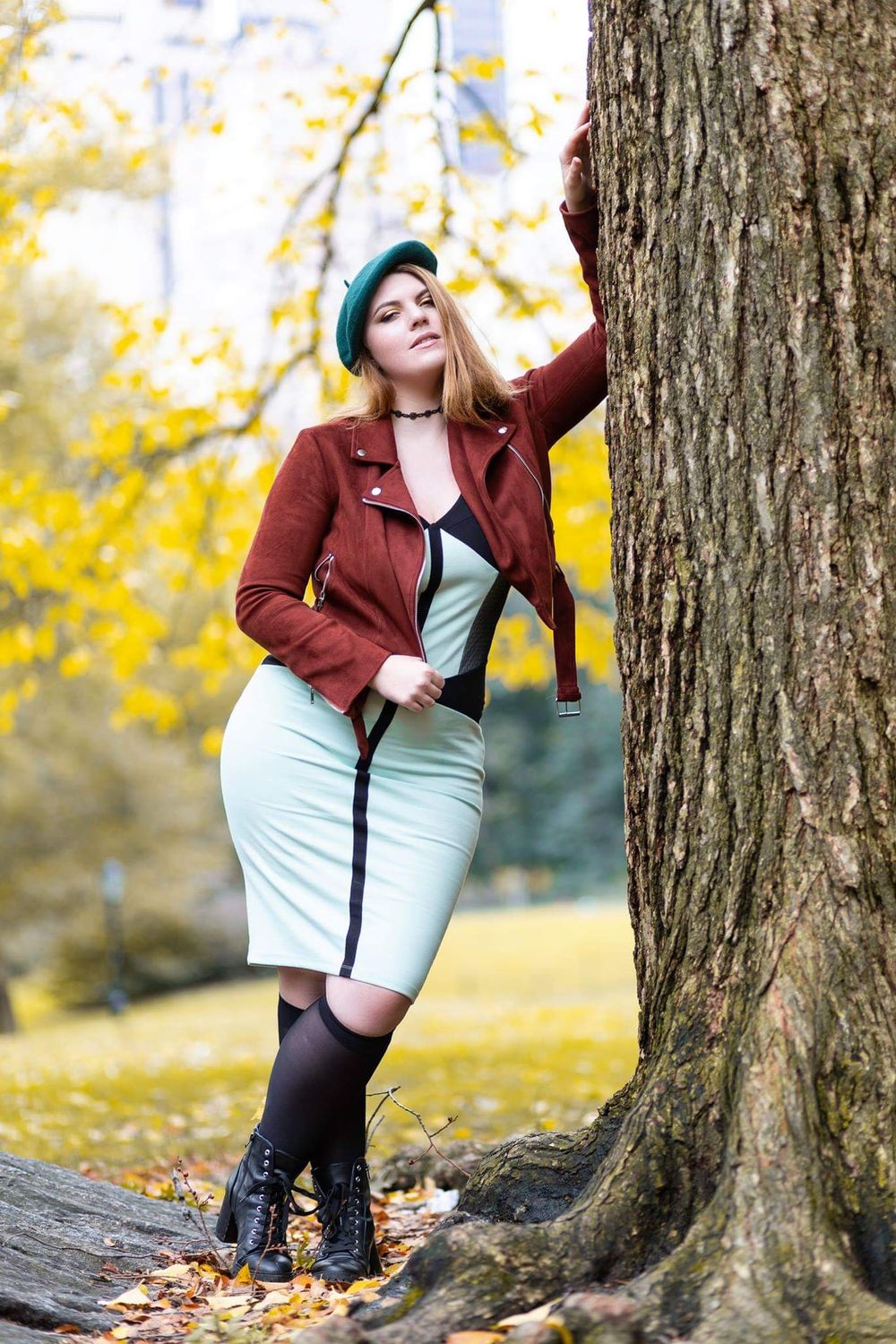 A person in a burgundy leather jacket and light dress poses playfully against autumn trees with golden foliage.