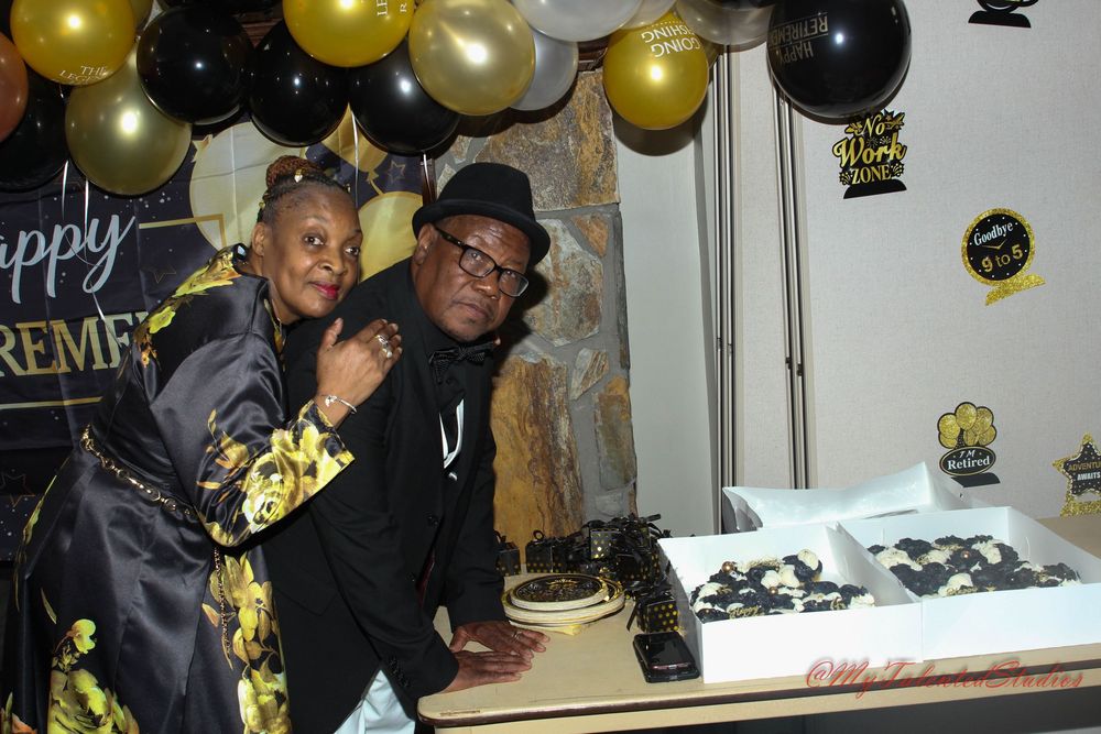 Couple poses together at retirement party beside cake under black and gold balloon arch.
