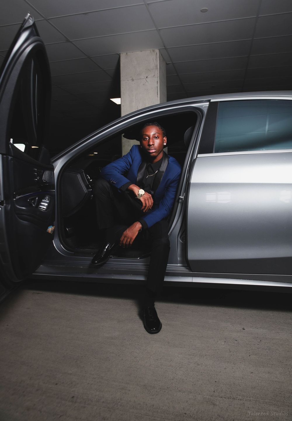 A stylish young man in a blue blazer sits in the open door of a silver luxury car.