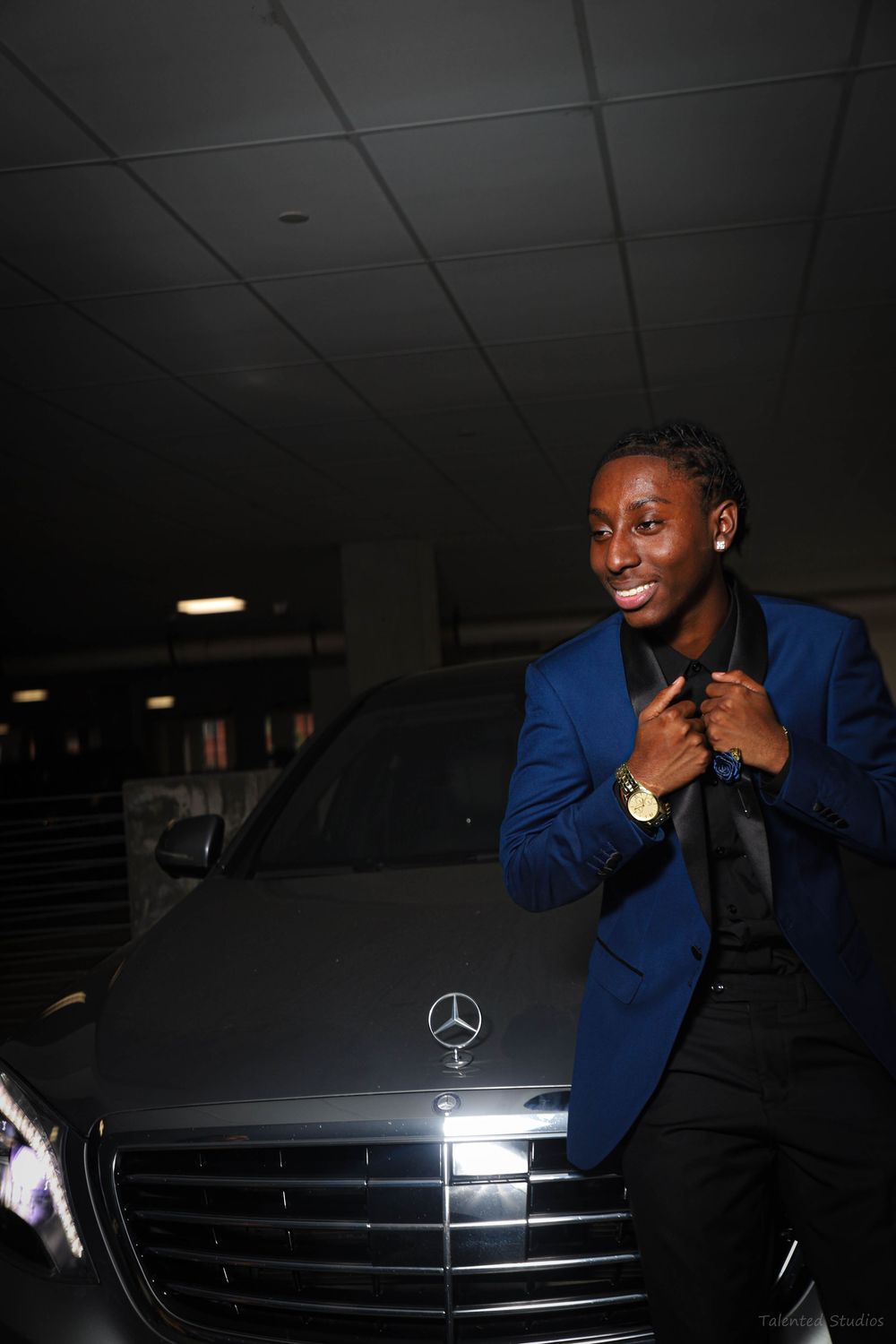 A young man in a blue blazer poses confidently beside a Mercedes-Benz in a parking garage.