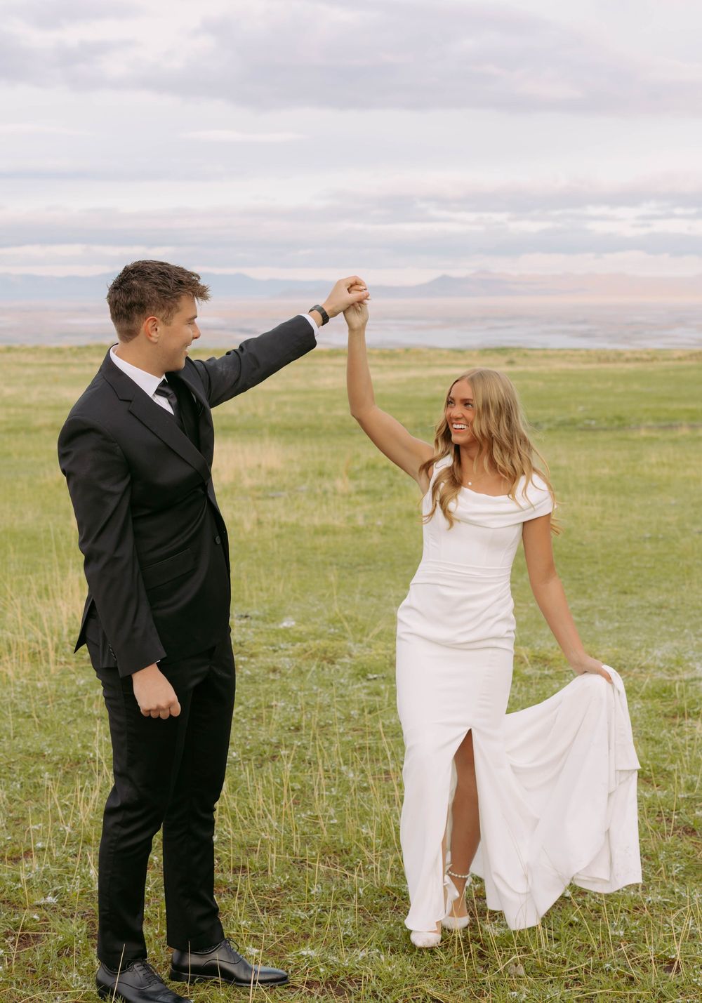 A couple in wedding attire dance together in a green field with mountains in the background at sunset.