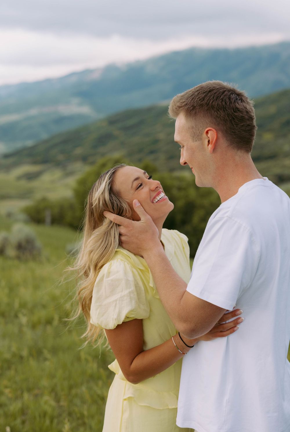 A romantic couple embraces and laughs together on a grassy hillside with mountains in the background.