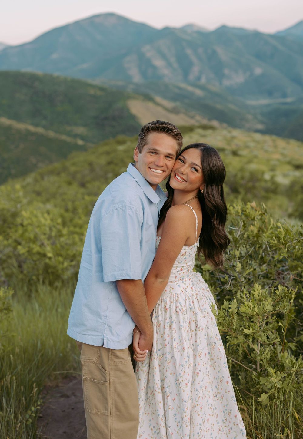 A couple poses together on a mountain trail with scenic green hills and mountains in the background at sunset.