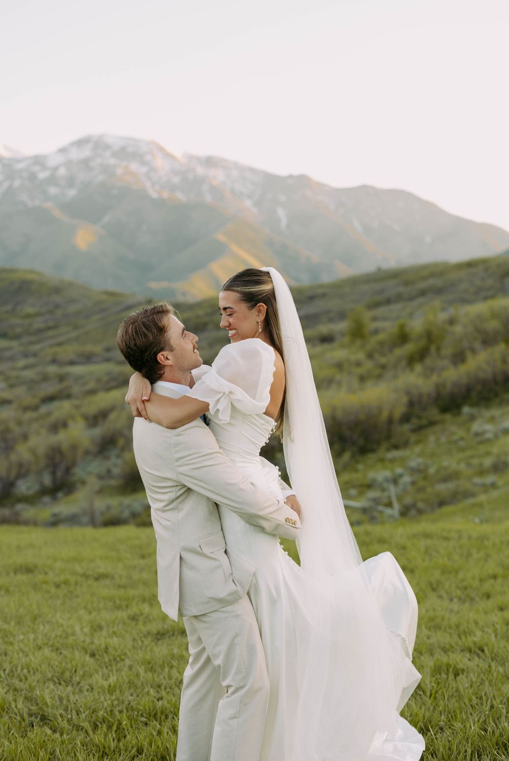 A newlywed couple embraces joyfully in a white wedding attire against a majestic mountain backdrop on green grass.