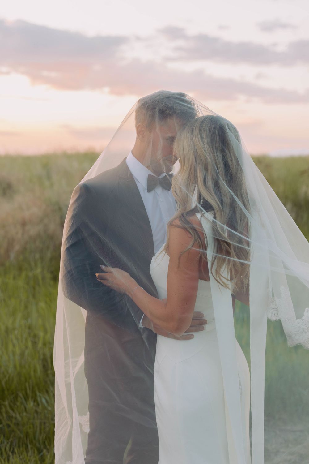 Newlywed couple embraces in field at sunset with bride's veil flowing in golden light.
