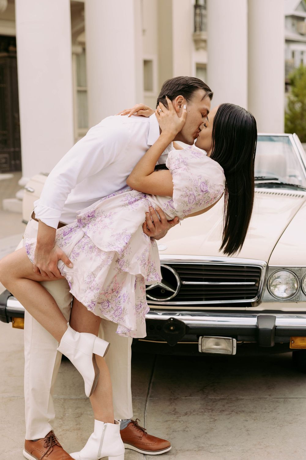 A romantic couple in vintage style clothing share an intimate dip kiss pose in front of a classic white Mercedes car.