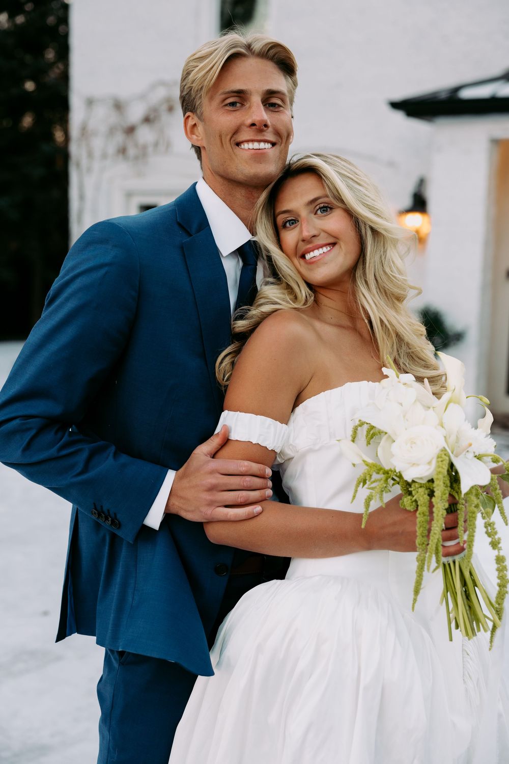 A happy couple on their wedding day, with the groom in a navy suit and bride in white holding a bouquet.