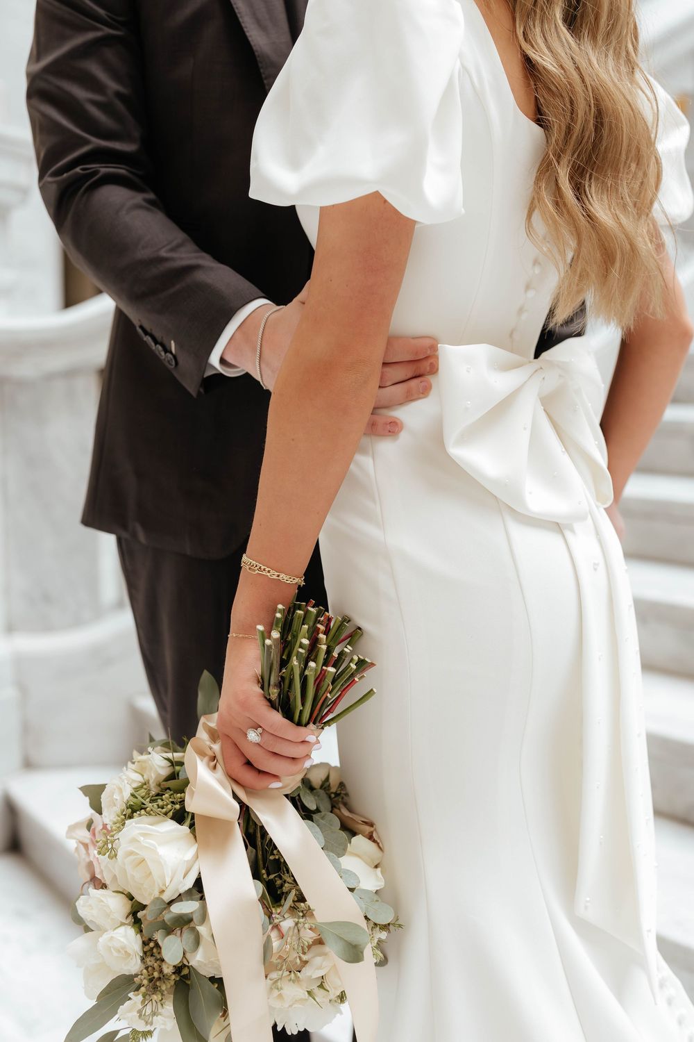 Close-up view of white bridal bouquet and elegant wedding dress details as couple stands together at ceremony.
