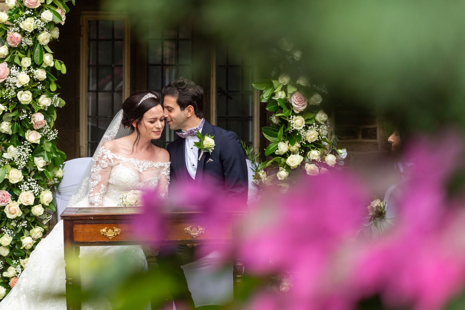 Romantic wedding portrait framed by pink flowers and greenery in a garden setting.