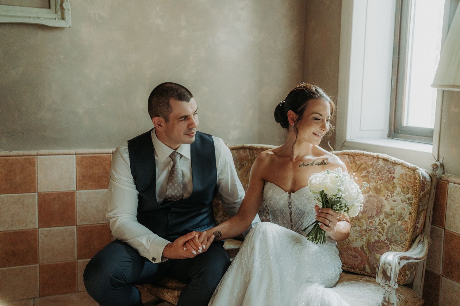 Bride in white coat holds autumn bouquet while groom stands behind near waterfront.