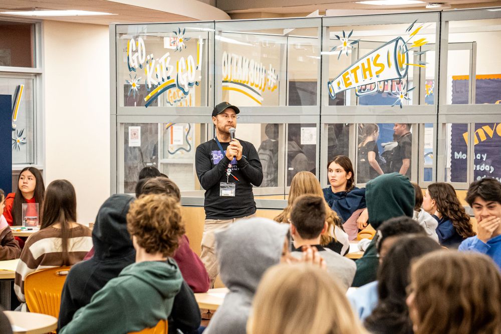 Students gather in a classroom listening attentively to someone speaking at the front near windows with decorative signage.