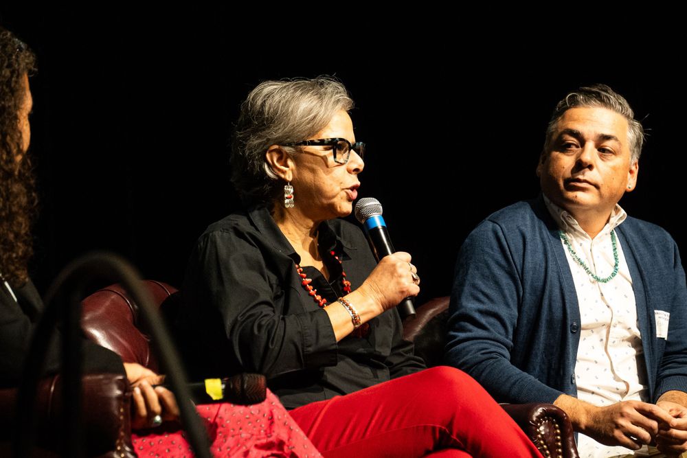 Two people sit during a panel discussion, with one speaking into a microphone while wearing bright red pants.