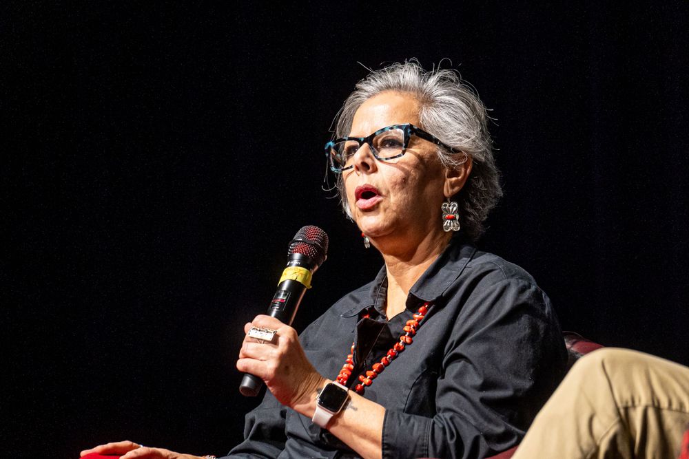 A speaker with gray hair and black glasses holds a microphone while speaking at a stage podium.