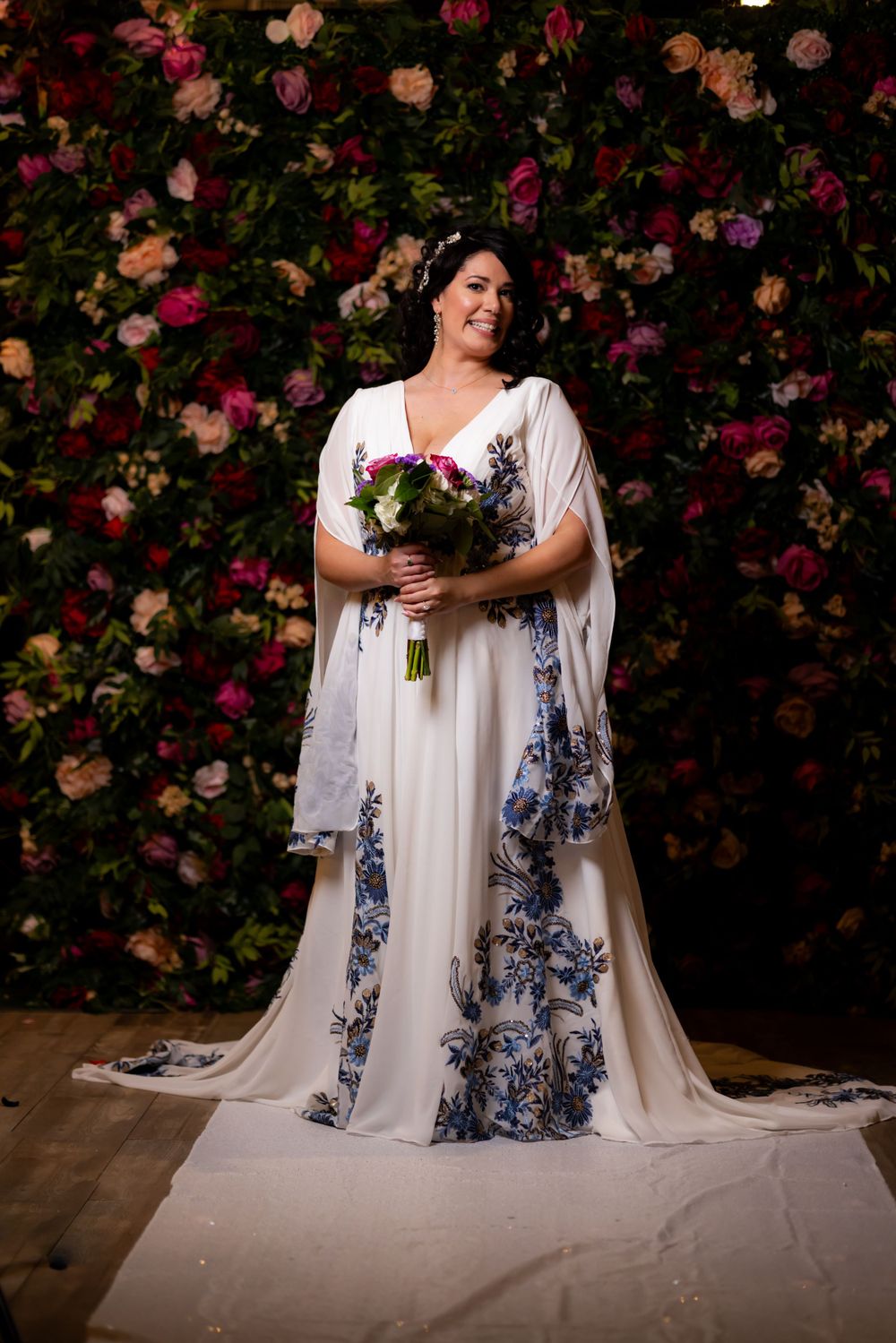 Bride in a white dress with blue floral details poses against a wall of pink and peach roses.