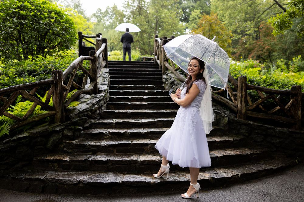 A bride in a white dress poses with an umbrella on stone steps in a lush Shakespeares Garden in Central Park NYCsetting.