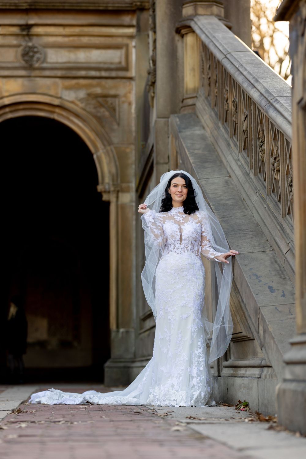 Elegant bride in lace wedding dress with flowing veil poses on historic stone staircase at Bethesda Fountain & Terrace.