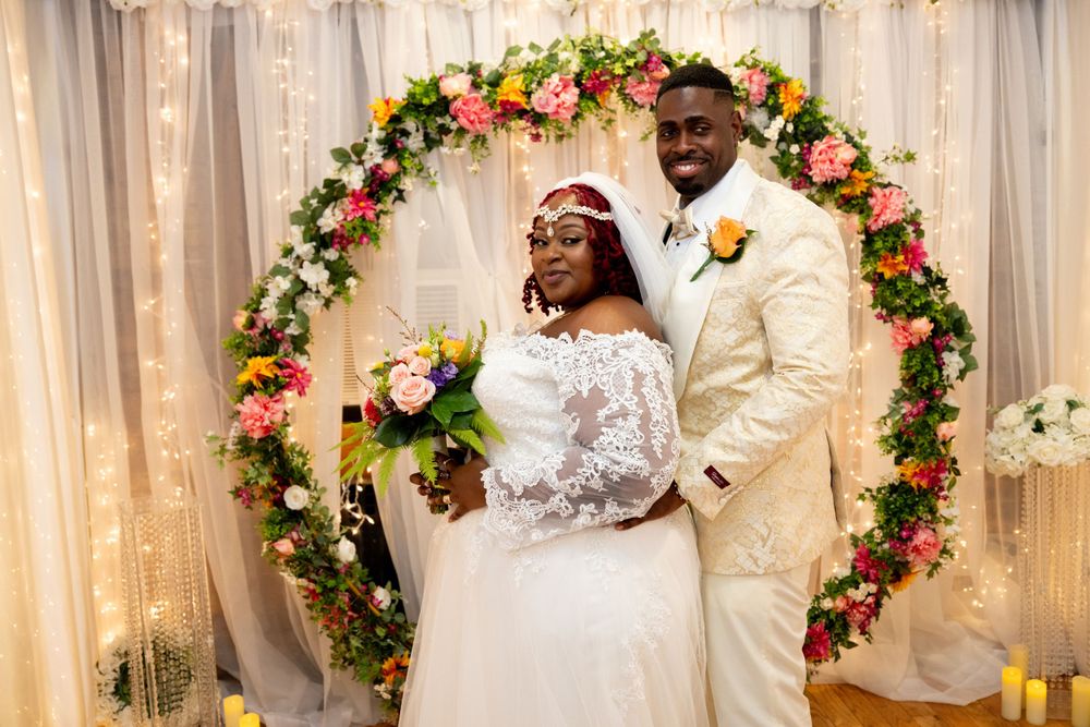 Wedding ceremony setup with floral arch featuring pink and white flowers against soft curtain backdrop.