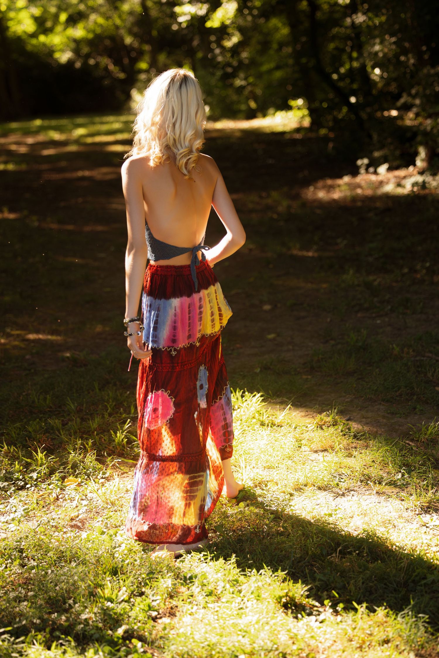 Woman in colorful tie-dye dress stands in sunlit forest clearing.