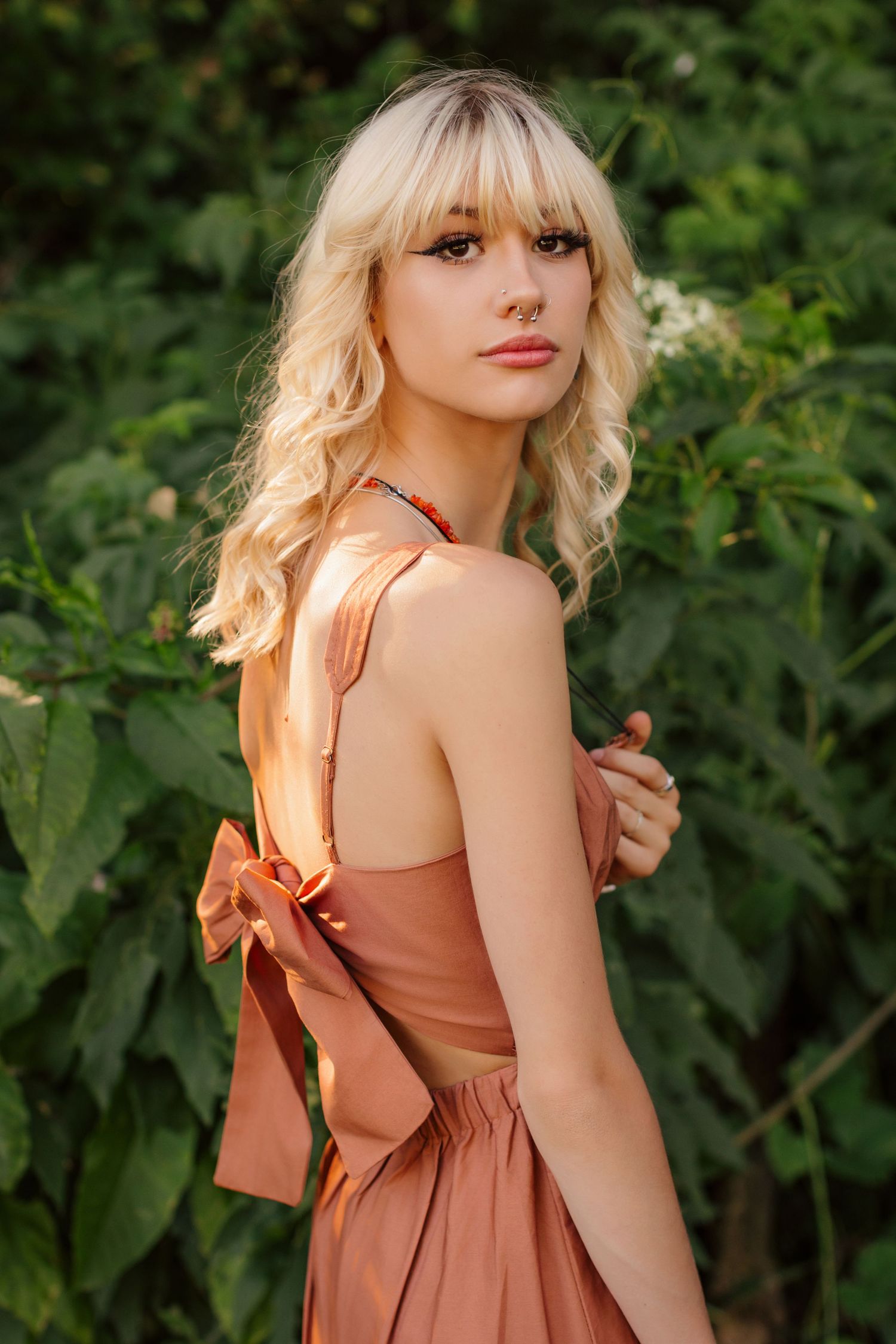 Portrait of woman in terracotta dress with bow detail against green foliage.