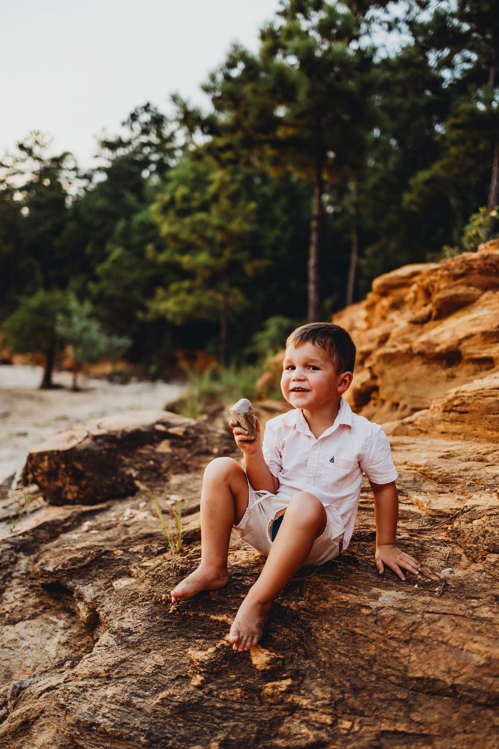 A child sits on orange-tinted rocks near a wooded beach at sunset.